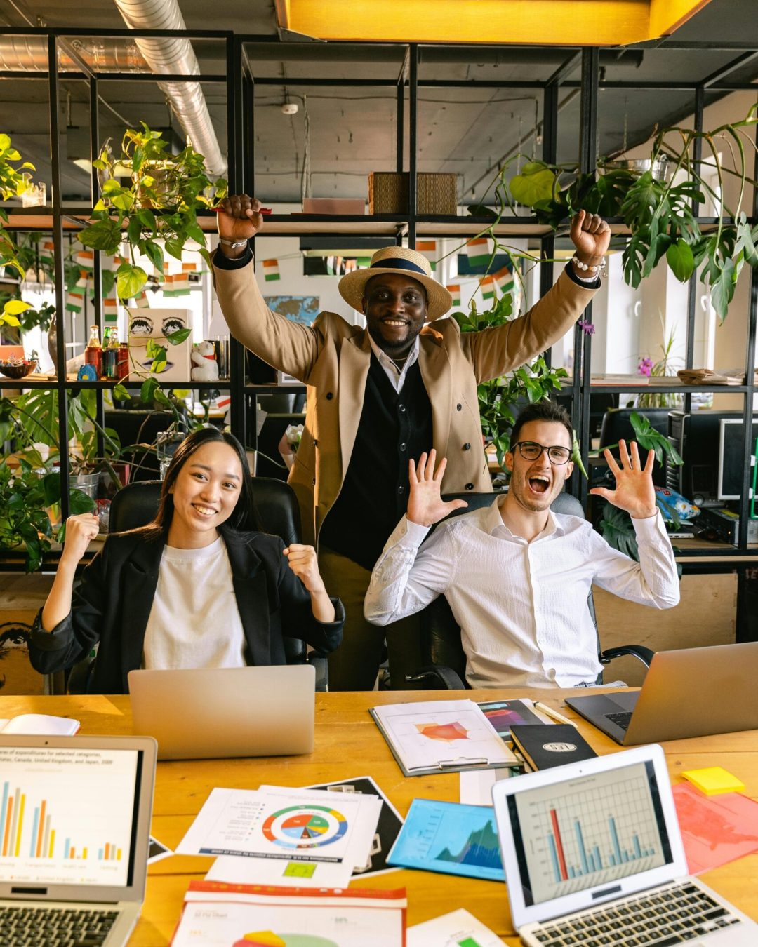 Happy diverse team celebrating success in a modern office with laptops and charts.